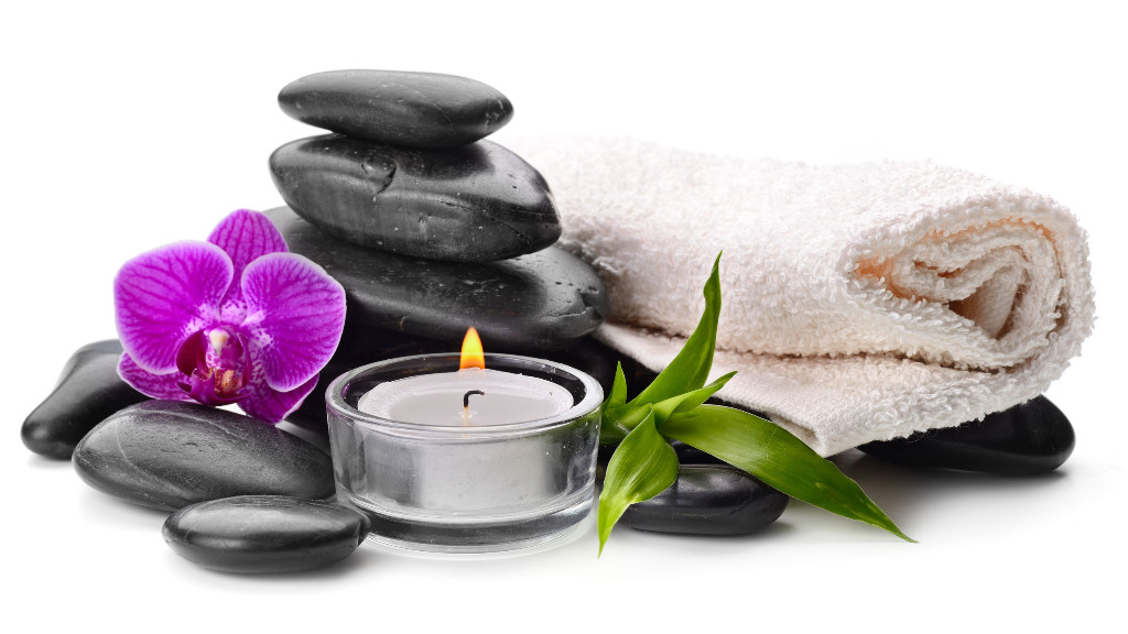 Das Bild zeigt Massage Steine, ein Handtuch, eine lila Orchidee sowie eine brennende Kerze